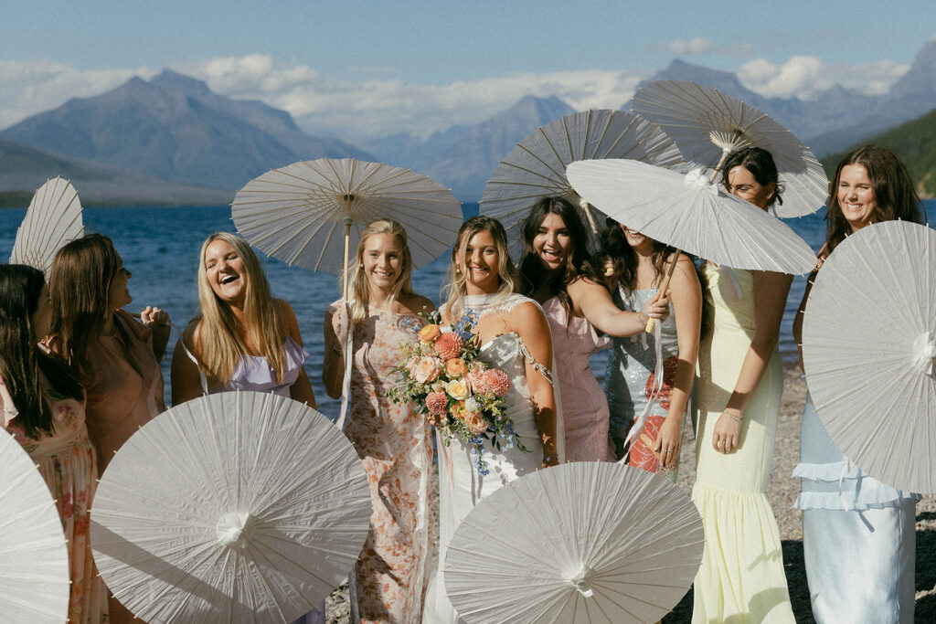 Bride and bridesmaids in colorful dresses on the shores of Lake McDonald with paper umbrellas.